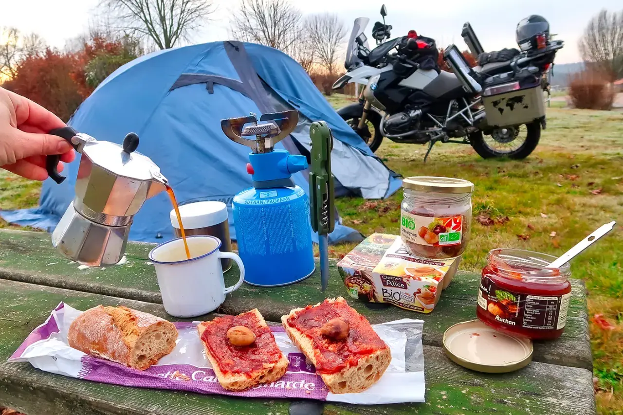 Colazione da campeggio in moto: caffè dalla moka, pane con marmellata e castagne, con tenda e moto sullo sfondo.