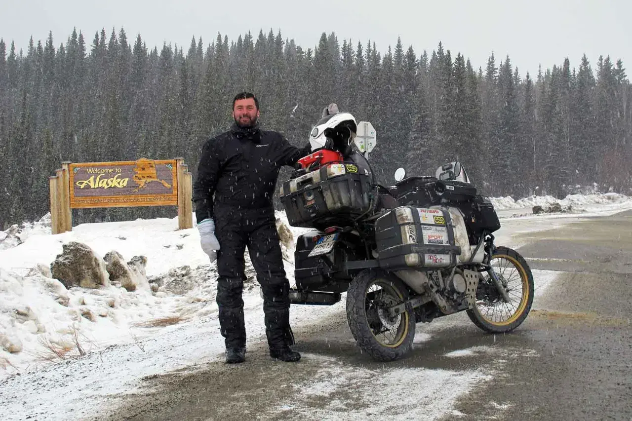 Un uomo in tuta da moto nera e guanti bianchi posa accanto alla sua moto carica, con un cartello "Welcome to Alaska" e una foresta innevata sullo sfondo. I suoi compagni di viaggio in moto sono pronti per l'avventura.