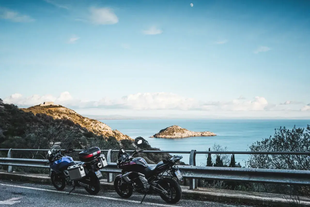 Due moto parcheggiate con vista sul mare e un'isola, con un rudere sulla collina. Un'avventura in **Argentario in moto** attende.