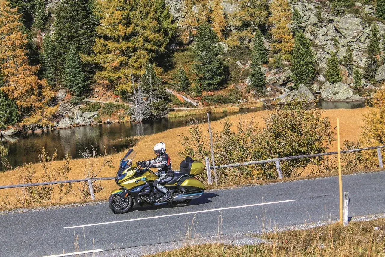 Motociclista in sella a una moto dorata affronta il **coccau valico** tra boschi autunnali e un lago alpino.
