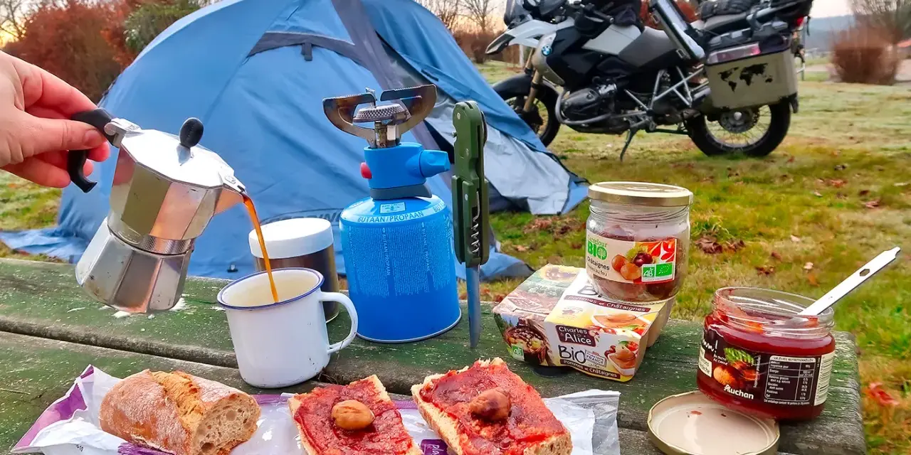 Colazione da campeggio in moto: caffè dalla moka, pane con marmellata e castagne, con tenda e moto sullo sfondo.