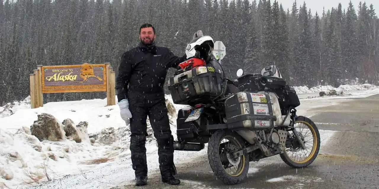Un uomo in tuta da moto nera e guanti bianchi posa accanto alla sua moto carica, con un cartello "Welcome to Alaska" e una foresta innevata sullo sfondo. I suoi compagni di viaggio in moto sono pronti per l'avventura.