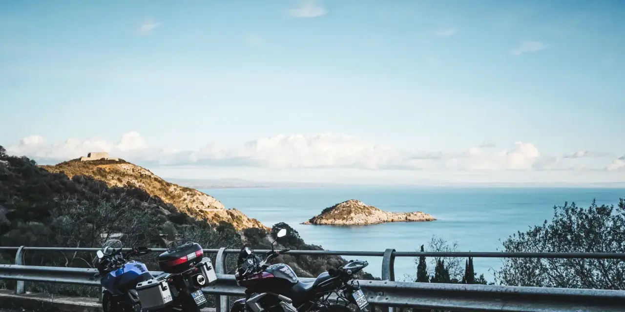 Due moto parcheggiate con vista sul mare e un'isola, con un rudere sulla collina. Un'avventura in **Argentario in moto** attende.