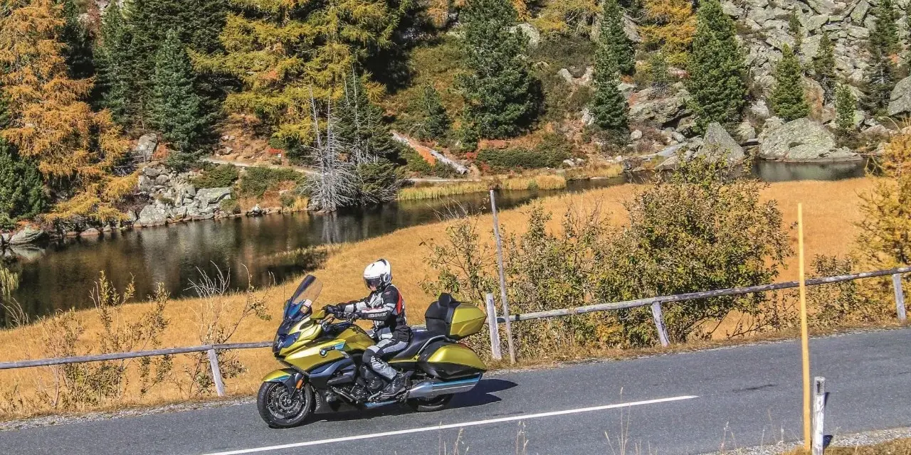 Motociclista in sella a una moto dorata affronta il **coccau valico** tra boschi autunnali e un lago alpino.