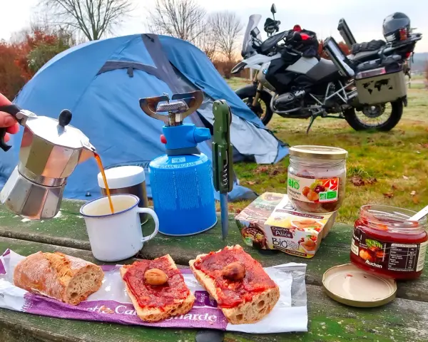 Colazione da campeggio in moto: caffè dalla moka, pane con marmellata e castagne, con tenda e moto sullo sfondo.