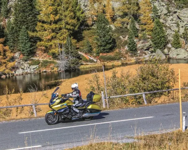 Motociclista in sella a una moto dorata affronta il **coccau valico** tra boschi autunnali e un lago alpino.