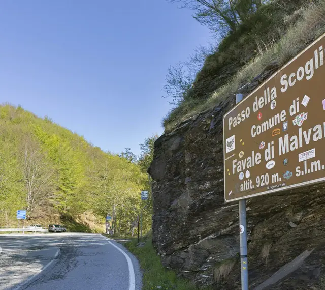 Cartello stradale indica "Passo della Scoglina, Favale di Malvaro, altit. 920 m. s.l.m." su una strada di montagna con vegetazione lussureggiante.