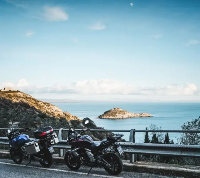 Due moto parcheggiate con vista sul mare e un'isola, con un rudere sulla collina. Un'avventura in **Argentario in moto** attende.