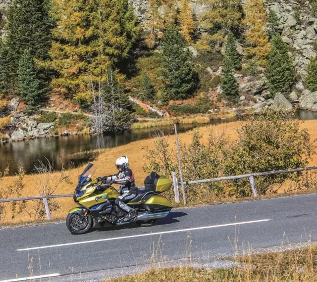 Motociclista in sella a una moto dorata affronta il **coccau valico** tra boschi autunnali e un lago alpino.