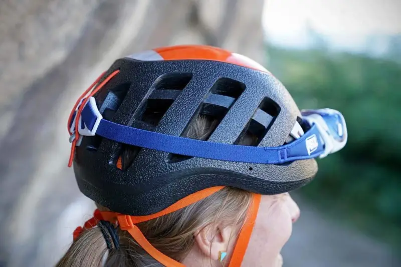 Casco da arrampicata nero con dettagli arancioni e lampada frontale. La cinghia blu aiuta ad allargare il casco stretto per un comfort ottimale.